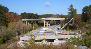 An on-ramp or off-ramp that is under construction. It has a concrete frame with metal beams sticking out of the sides, but is not yet paved. It's going under a bridge with forest on either side.