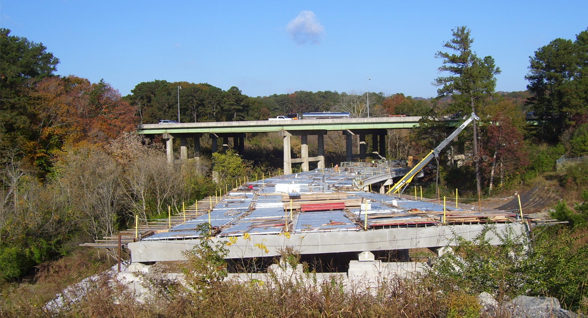 An on-ramp or off-ramp that is under construction. It has a concrete frame with metal beams sticking out of the sides, but is not yet paved. It's going under a bridge with forest on either side.