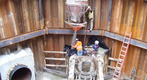 Construction workers pour concrete into a column, in front of a segmented wooden wall.