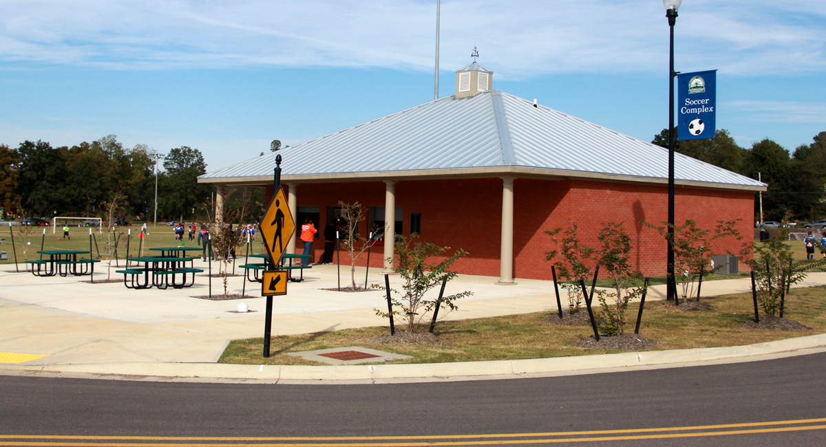 A pavilion in a park. There are picnic tables in front and a soccer field in the background.