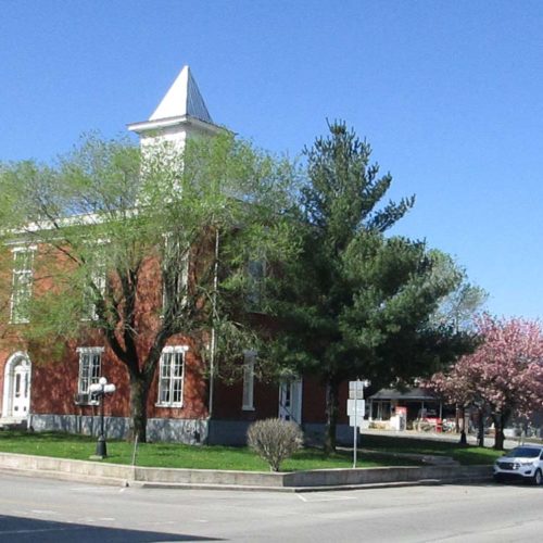 A rectangular brick building with a tower in the middle. On the right side of the frame is a Celina Walks logo: a black silhouette shape of the building with a pedestrian walking sign in the middle.