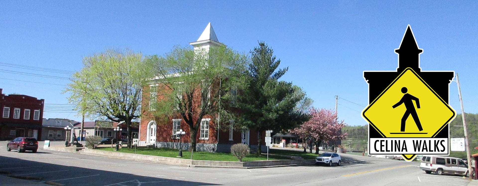 A rectangular brick building with a tower in the middle. On the right side of the frame is a Celina Walks logo: a black silhouette shape of the building with a pedestrian walking sign in the middle.