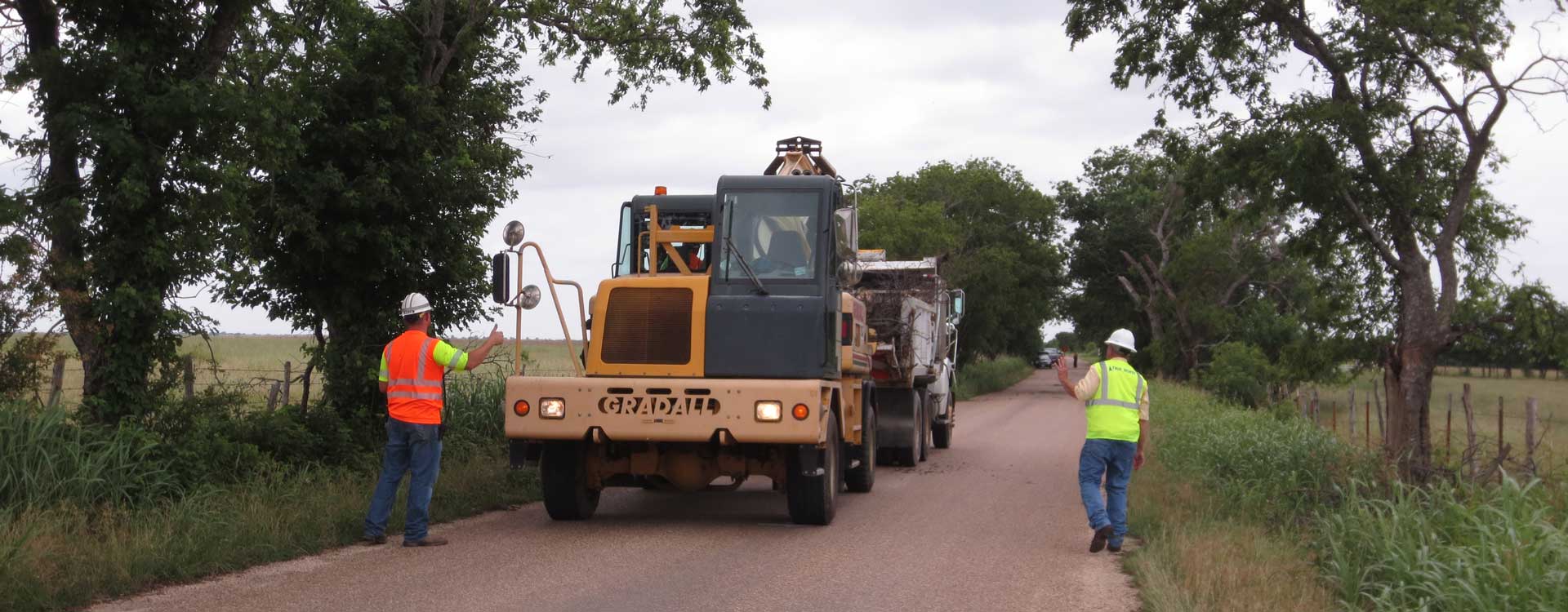 Two construction workers directing an excavator on an unpaved road.