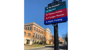 A sign on a road in front of a brick building. It has arrows pointing towards "Tax Assessor," "City Hall," "Visitors Center," "Saenger Theater," and "Public Parking."