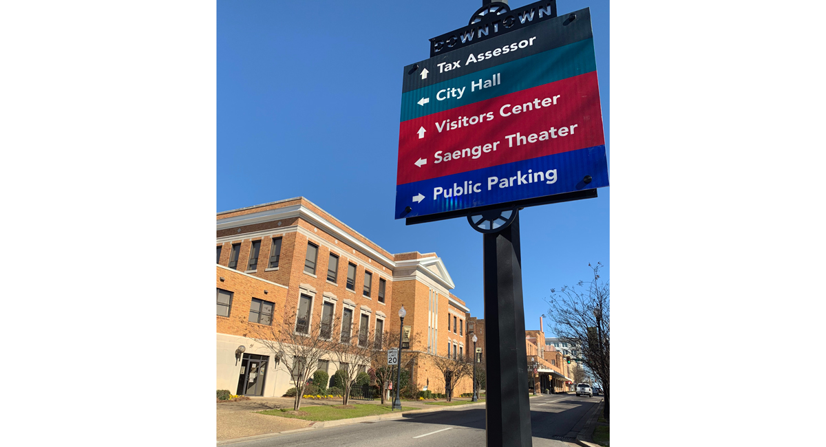 A sign on a road in front of a brick building. It has arrows pointing towards "Tax Assessor," "City Hall," "Visitors Center," "Saenger Theater," and "Public Parking."