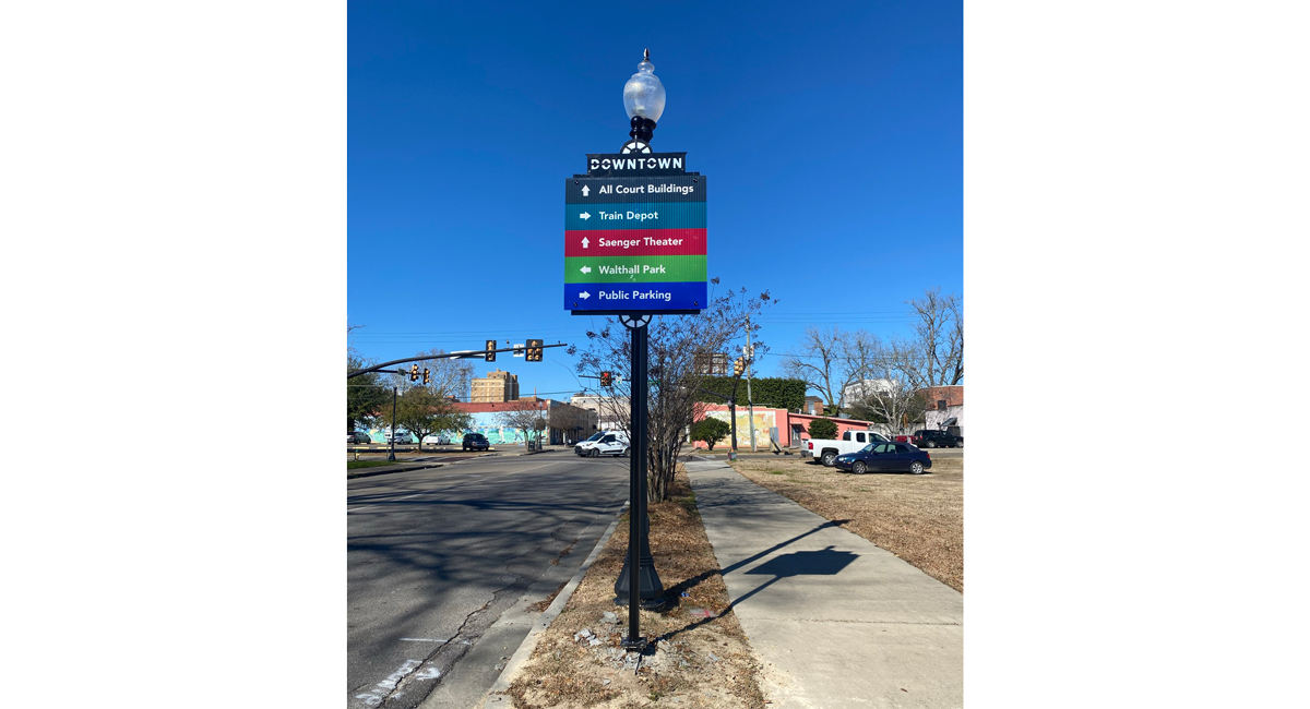 A sign next to a sidewalk in front of an intersection. It gives directions to "All Court Buildings," "Train Depot," "Saenger Theater," "Walthall Park," and "Public Parking."